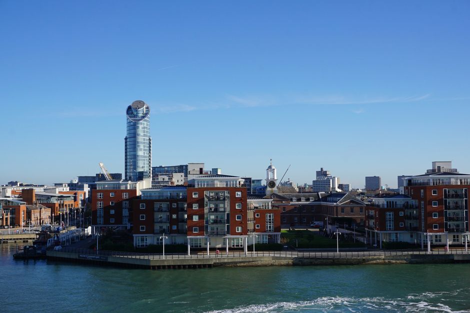 A panoramic daytime view of a modern urban waterfront in Chelsea, showing a cluster of red-brick residential buildings with balconies positioned along the water's edge, with several white-finished boats docked nearby. Behind the buildings, there is a tall, distinctive glass skyscraper with a rounded top, and various other mid-rise structures forming the city skyline under a clear blue sky. This scene is relevant to house removals and furniture transport, illustrating the type of environment where professional removals company Chelsea may operate, including moving logistics and packing considerations for apartments close to the river, as discussed in the Chelsea Harbour apartment removals and lift access advice page.