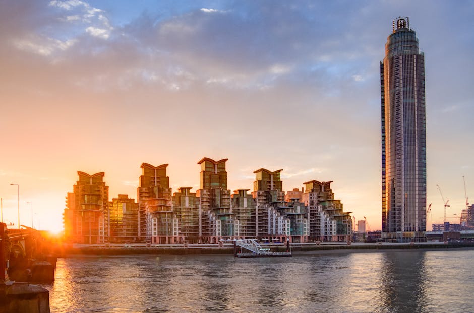 A panoramic view of Chelsea Harbour in the early evening, featuring a cluster of modern residential buildings with distinctive rooftop designs situated on the waterfront. The buildings are constructed with a combination of glass, concrete, and steel, reflecting warm sunset light. To the right, a tall, cylindrical skyscraper rises prominently, with reflective glass windows and a rounded top. The scene includes a river in the foreground with gentle ripples, and a boat docked near the riverbank. Alongside, construction cranes are visible in the background, indicating ongoing development. The setting sun casts a soft glow over the scene, with partly cloudy sky enhancing the urban landscape. This image showcases the contemporary architecture and skyline of Chelsea Harbour, relevant for house removals and relocation services offered by Removal Companies Chelsea, especially when considering logistics involving high-rise or waterfront properties and lift access advice.
