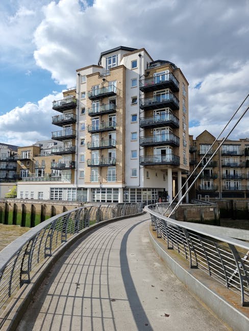 A panoramic daytime view of a modern urban waterfront in Chelsea, showing a cluster of red-brick residential buildings with balconies positioned along the water's edge, with several white-finished boats docked nearby. Behind the buildings, there is a tall, distinctive glass skyscraper with a rounded top, and various other mid-rise structures forming the city skyline under a clear blue sky. This scene is relevant to house removals and furniture transport, illustrating the type of environment where professional removals company Chelsea may operate, including moving logistics and packing considerations for apartments close to the river, as discussed in the Chelsea Harbour apartment removals and lift access advice page.