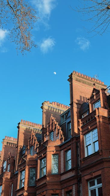 A clear daytime view of the facades of narrow brick terrace houses on Kings Road in Chelsea, featuring ornate architectural details, tall windows with white frames, and decorative crenellations along the roofline. The sky above is bright blue with minimal cloud cover, and a small section of tree branches without leaves is visible at the top right corner. In the context of house removals, the image captures the urban environment where furniture and boxes, possibly wrapped in plastic or cardboard, could be loaded onto a van by Removal Companies Chelsea during a home relocation process, with a focus on loading and transport logistics for narrow Chelsea terraces.