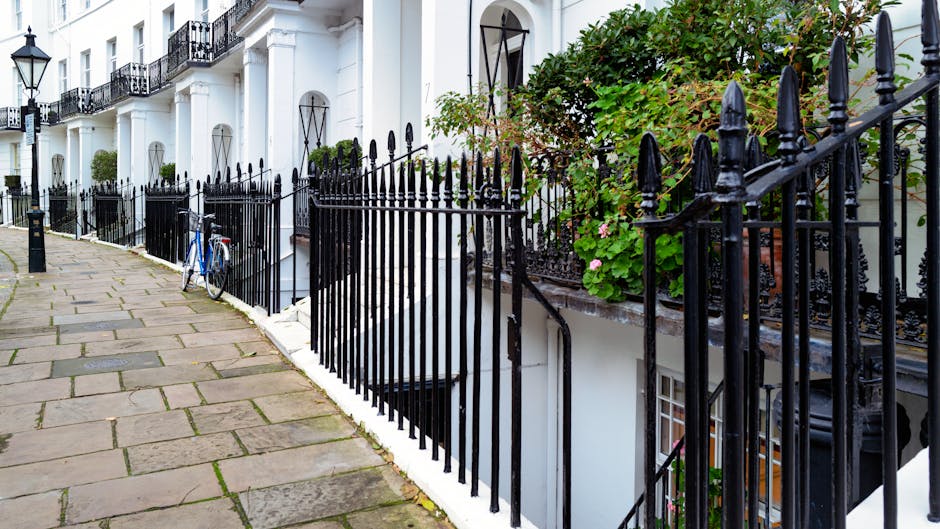 Image showing a narrow cobblestone pavement running alongside white terraced houses with black wrought iron railings and gates. A blue bicycle is parked against the fence, and a small patch of greenery with pink flowers is visible above the railing. The front entrances of the houses feature large windows and black ironwork details, typical of Chelsea terrace architecture. The scene is well-lit with natural daylight, and the overall environment reflects a quiet residential street suitable for house removals and furniture transport services, as documented on the Kings Road removals guide for narrow Chelsea terraces provided by Removal Companies Chelsea.