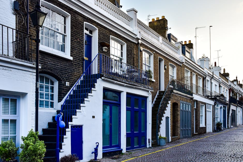 A clear daytime view of the facades of narrow brick terrace houses on Kings Road in Chelsea, featuring ornate architectural details, tall windows with white frames, and decorative crenellations along the roofline. The sky above is bright blue with minimal cloud cover, and a small section of tree branches without leaves is visible at the top right corner. In the context of house removals, the image captures the urban environment where furniture and boxes, possibly wrapped in plastic or cardboard, could be loaded onto a van by Removal Companies Chelsea during a home relocation process, with a focus on loading and transport logistics for narrow Chelsea terraces.