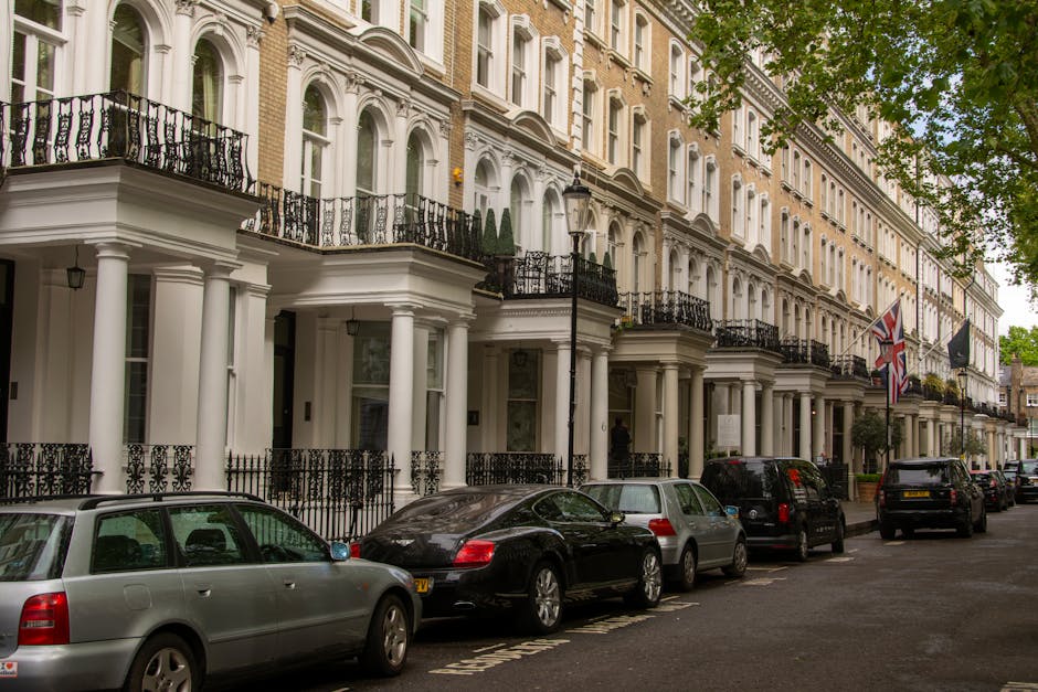 A row of elegant Victorian-style terraced houses in Sloane Square Chelsea, featuring white facades with decorative columns, black wrought-iron balconies, and large sash windows. The street is lined with parked cars, including a silver estate car, a black sedan, and other vehicles, positioned alongside a paved sidewalk with a lamppost and some greenery visible in the background. Flags, including the Union Jack, are displayed on some of the buildings. The scene is outdoors during the daytime with natural light illuminating the buildings and street, capturing the typical residential environment near Duke of York Square. This setting illustrates a typical location for home relocation or furniture transport services by Removal Companies Chelsea, highlighting the urban architecture and street layout associated with house removals near Chelsea.