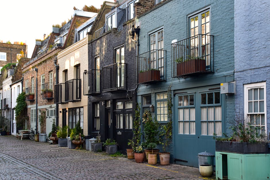 A row of terraced townhouses in Chelsea with cobbled street in front, featuring mixed exterior wall finishes in shades of blue, beige, and black. Each property has small front gardens or potted plants with various containers like terracotta, plastic, and metal pots. Some houses display small balconies with black metal railings and flower boxes. Large windows with divided panes are visible, alongside front doors painted in coordinating colours. The scene is illuminated by natural daylight, highlighting the textures of the brick and plaster facades. This image reflects the typical urban residential setting where house removals and furniture transport may occur, with visible features suitable for professional relocation and packing services by companies such as Removal Companies Chelsea, supporting house moving logistics in Chelsea mews and townhouses.