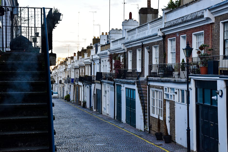 A row of terraced houses in Chelsea, London, featuring brick facades with white window frames and decorative railings on small front balconies. The street is paved with cobblestones and the scene is illuminated by natural daylight, with the sky visible in the background. The houses have garage doors on the ground level, some painted in dark green or blue, and potted plants can be seen on a few balconies. On the left side of the image, a black metal staircase with steps leading upwards is partially visible. This setting reflects a typical residential area suitable for house removals or home relocation services, coordinated by companies such as Removal Companies Chelsea, who manage furniture transport and packing logistics in the area.